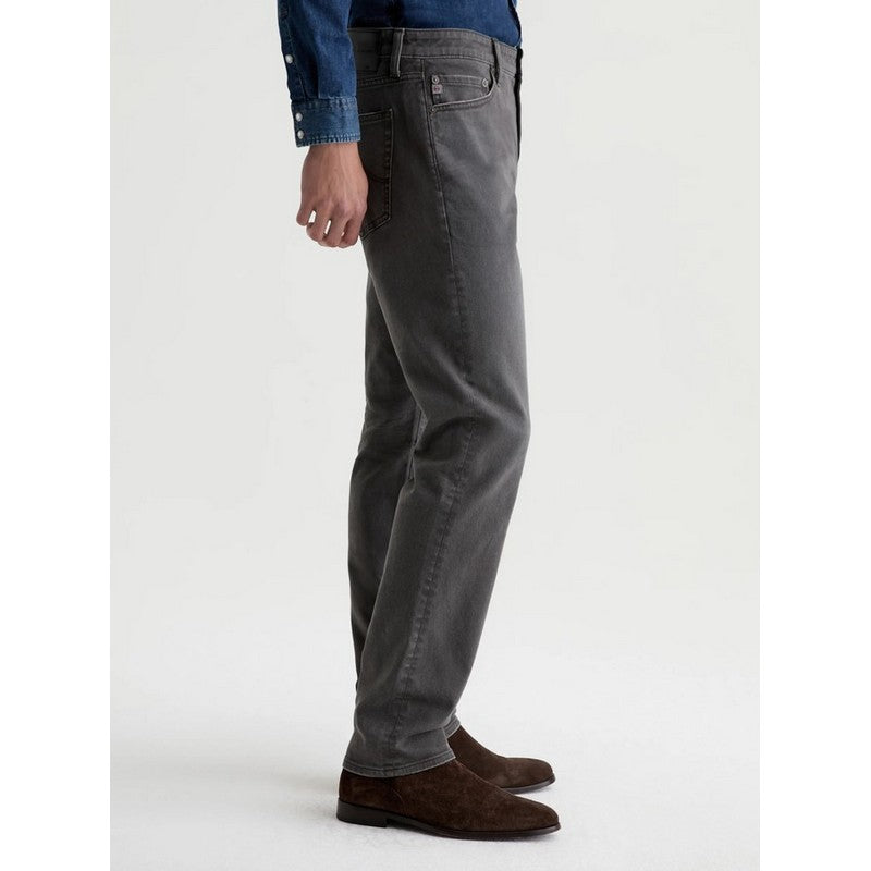 A person shown from the waist down wearing dark gray slim-fit jeans, a blue denim shirt, and brown suede shoes, standing against a plain light gray studio background.