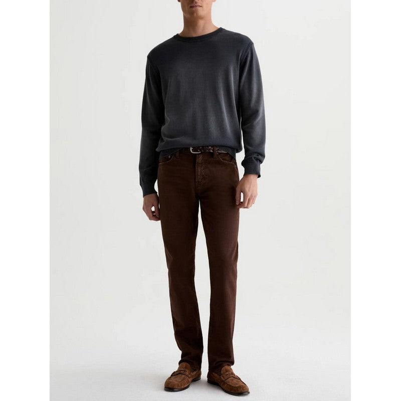 A man wearing a dark charcoal gray long-sleeve crewneck sweater, brown trousers, and brown suede loafers, standing against a plain light gray studio background.
