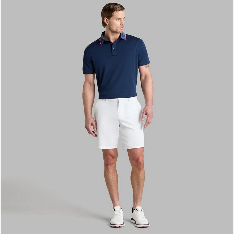 A man modeling a navy blue short-sleeve polo shirt with a contrasting red and blue inner collar, paired with white golf shorts and white golf shoes, against a solid gray background.