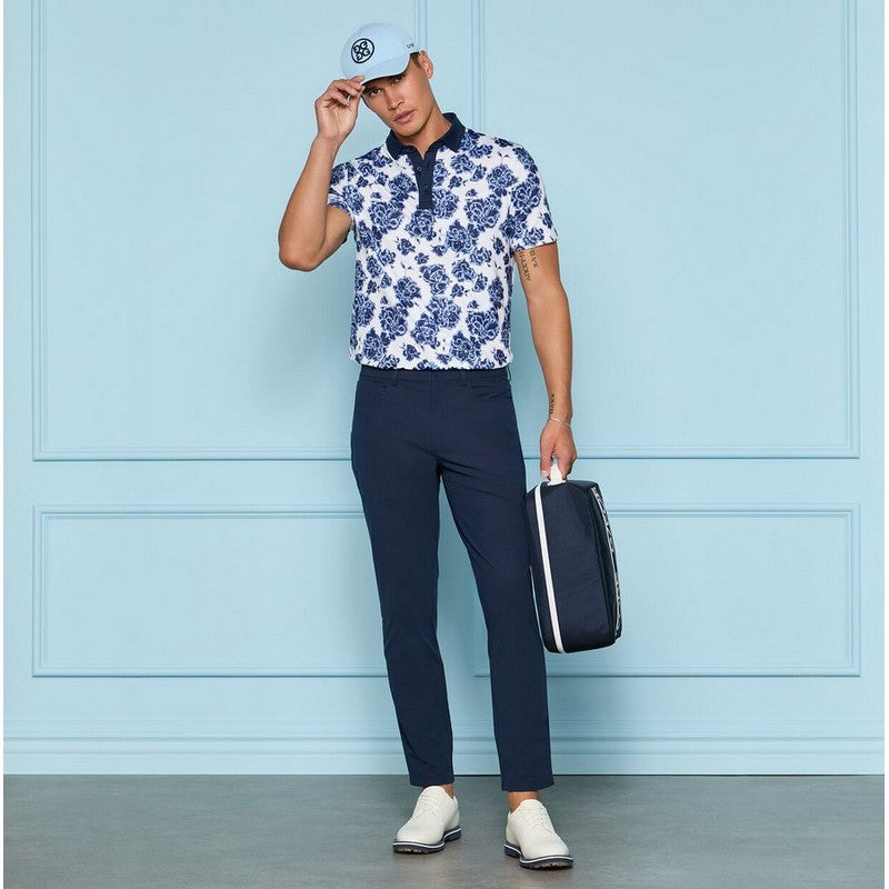 Person wearing a blue and white floral shirt with navy pants, holding a golf bag against a light blue wall.