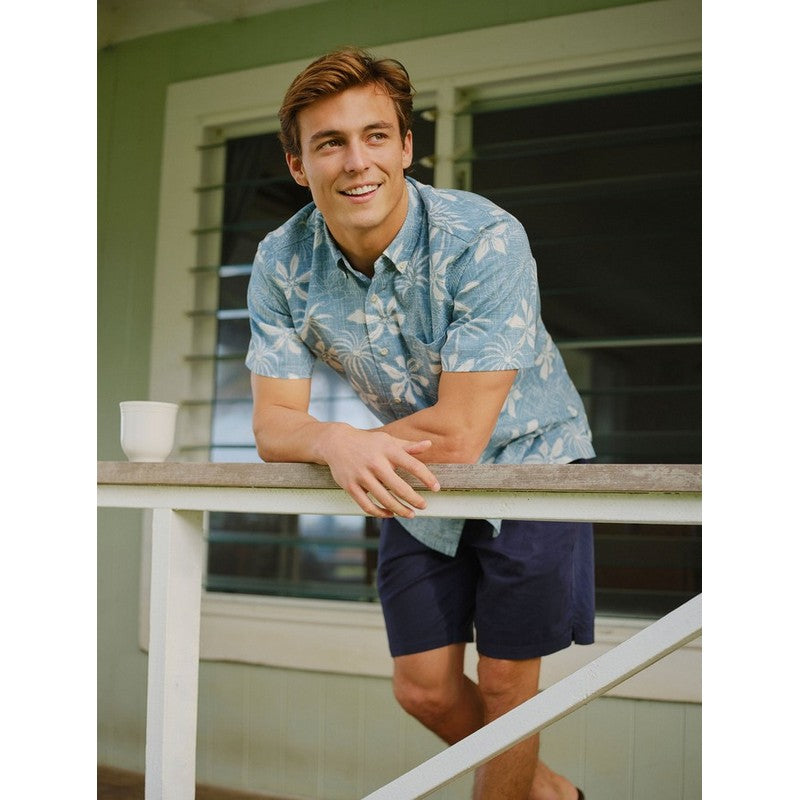 Man in a blue floral shirt and navy shorts leaning on a railing outdoors.